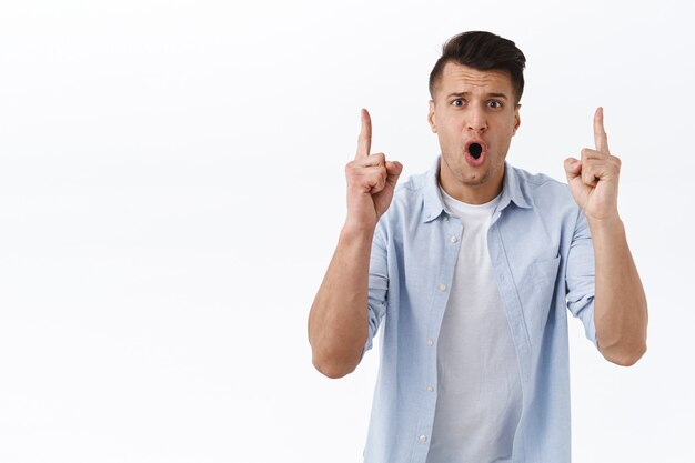 Portrait of alarmed and thrilled young concerned man showing something disturbing, pointing fingers up telling you urgent shocking news, standing astounded over white wall