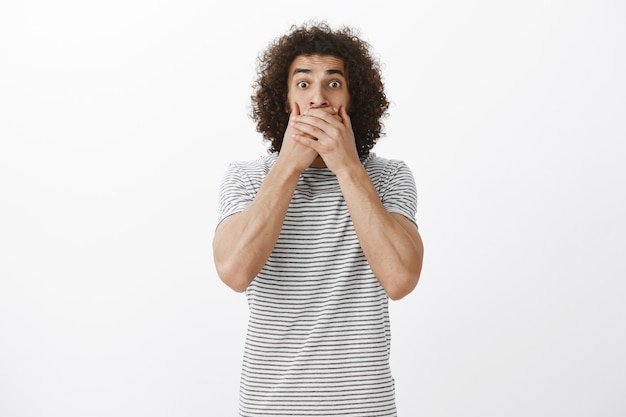 Free photo portrait of amazed anxious hispanic male model with afro hairstyle, staring with popped eyes and covering mouth with palms not to scream, being shocked and scared