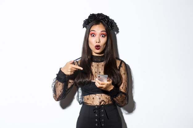 Portrait of amazed asian girl pointing finger at smartphone, showing something about halloween in internet, standing over white background in gothic lace dress.