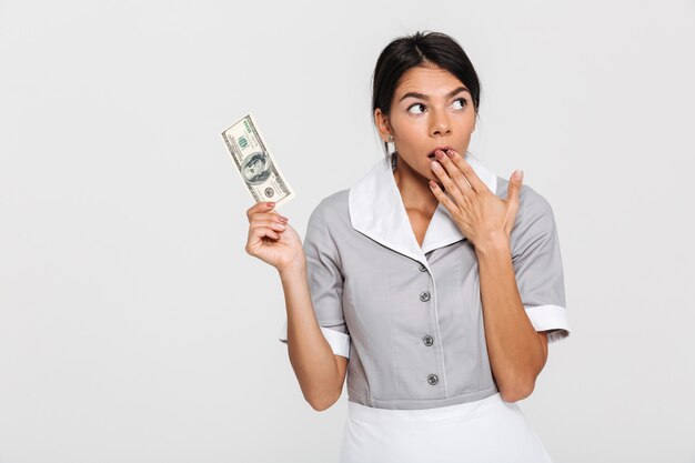 Portrait of amazed attractive maid in uniform holding hundred dollars while covering her mouth with hand, looking aside