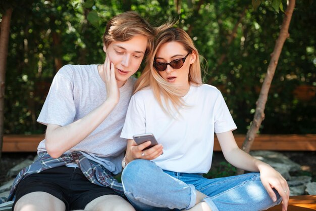 Ritratto di coppia stupita seduta su una panchina nel parco e guardando premurosamente nel cellulare foto ravvicinata di giovane uomo e bella signora con i capelli biondi che trascorrono del tempo insieme mentre si utilizza il telefono cellulare