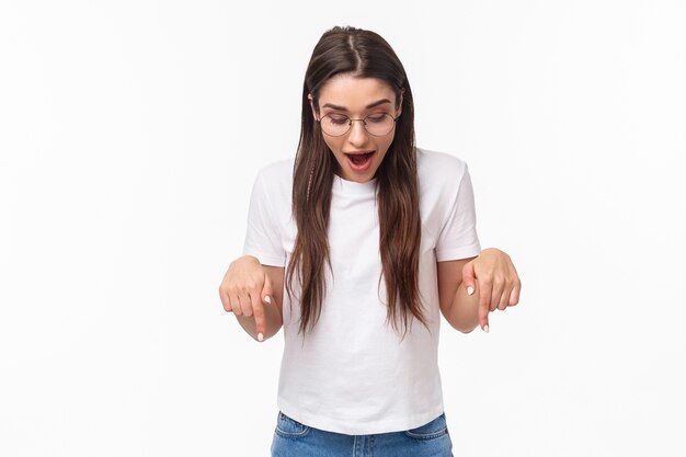 Portrait of amazed, curious happy brunette woman in t-shirt, glasses, looking and pointing down