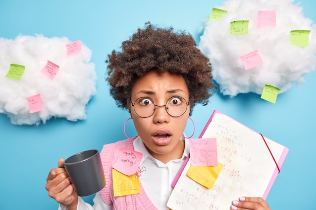 Free photo portrait of amazed curly haired woman drinks coffee busy doing startup project holds folder with papers surrounded by post it stickers nite to remember everything wears round spectacles poses in office