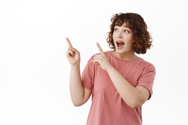 Portrait of amazed and excited beautiful girl, gasping and saying wow, pointing and looking at upper left corner copyspace, showing something amazing, standing against white background