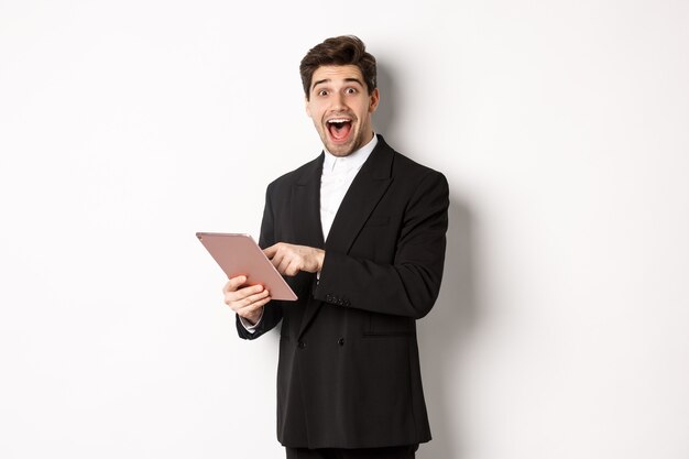 Портрет удивленного красивого бизнесмена в модном костюме, показывающего что-то классное на цифровом планшете, стоящем на белом фоне.