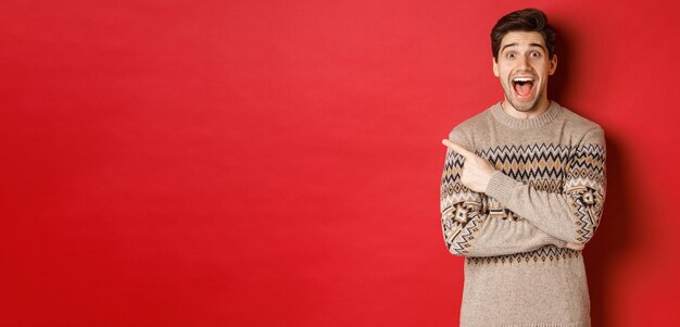 Portrait of amazed handsome guy wearing christmas sweater pointing finger at upper left corner logo and looking excited showing awesome new year promo offer standing over red background