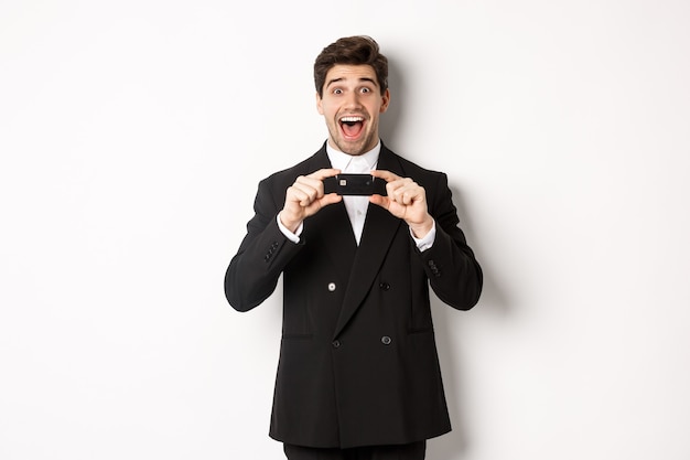 Portrait of amazed handsome man in black suit, showing credit card and recommending bank, standing over white background