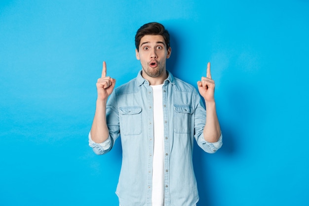 Portrait of amazed handsome man pointing fingers up, showing logo on blue background