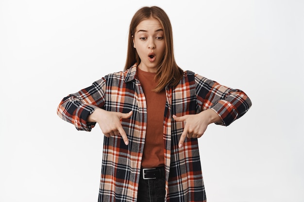 Free photo portrait of amazed happy woman, open mouth say wow surprised, pointing down and staring at banner, looking awesome logo, big discounts, white wall