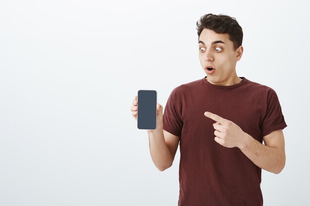 Портрет удивленного впечатленного красивого парня в красной футболке, показывающего новый смартфон