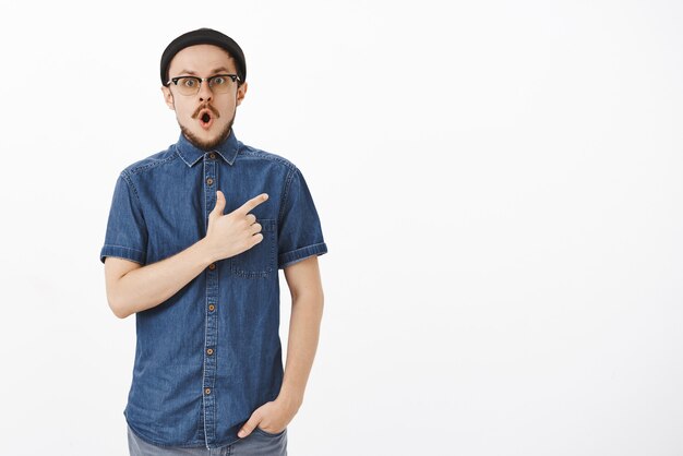 Portrait of amazed and impressed handsome young bearded guy in stylish beanie and blue shirt dropping jaw in wow sound pointing at upper right corner staring questioned and thrilled over white wall