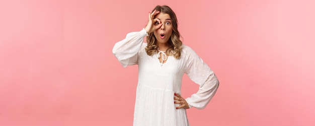 Portrait of amazed and intrigued young blond girl seeing something awesome look from okay sign with startled excited expression fold lips say wow pop eyes at camera pink background