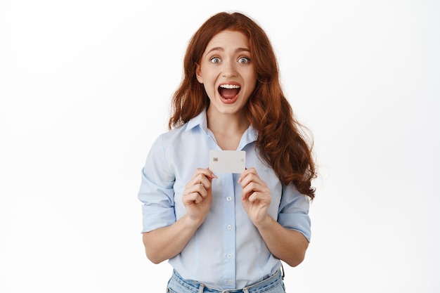 Free photo portrait of amazed redhead woman bank client, showing credit card and gasping astonished, telling about discounts, contactless payment, banking promotion, standing against white background.