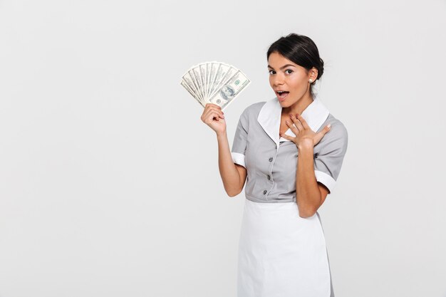 Portrait of amazed young housekeeper in uniform holding fan of dollar banknotes