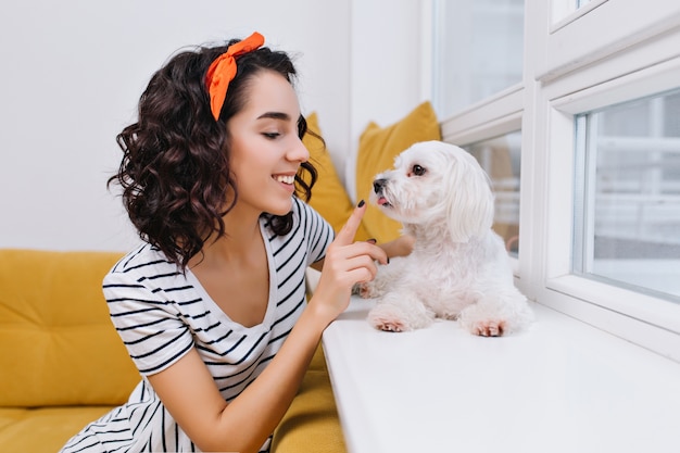 近代的なアパートで小さな犬と遊んで驚くべきうれしそうなファッショナブルな若い女性の肖像画。家庭でのペット、笑顔、陽気な気分、家で楽しむ