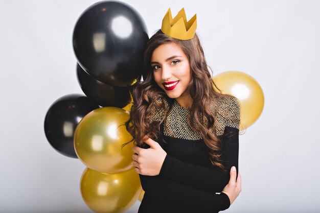 Portrait amazing pretty girl with long curly brunette hair, yellow crown,  black and gold balloons on white space.