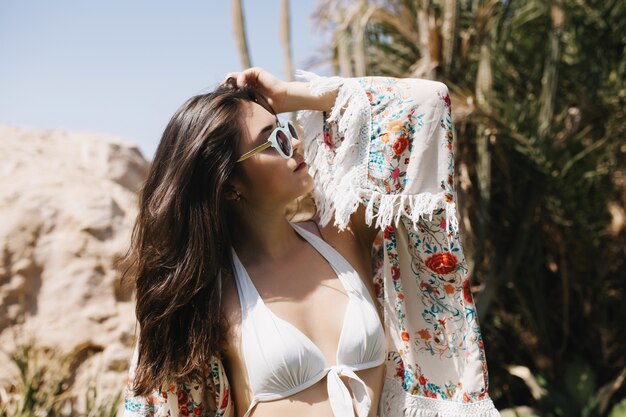 熱帯の国で休暇を楽しんでいるトレンディなサングラスで驚くほどスリムなブルネットの少女の肖像画。白い水着で優雅な若い女性がビーチで日光浴