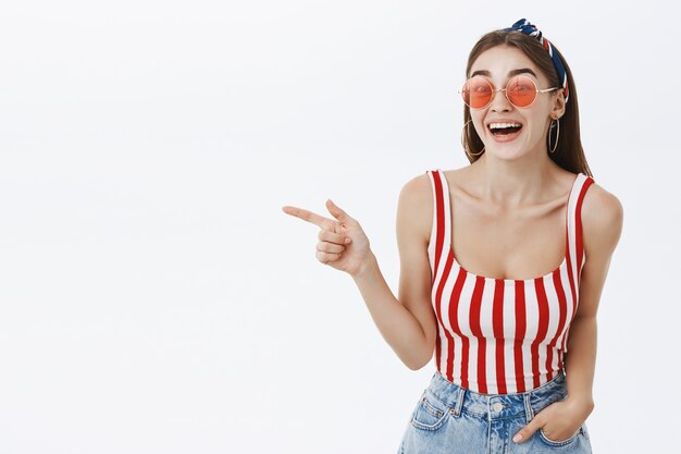 Portrait of amused good-looking stylish woman in striped top and sunglasses laughing out loud having fun pointing left at funny interesting copy space gazing joyfully