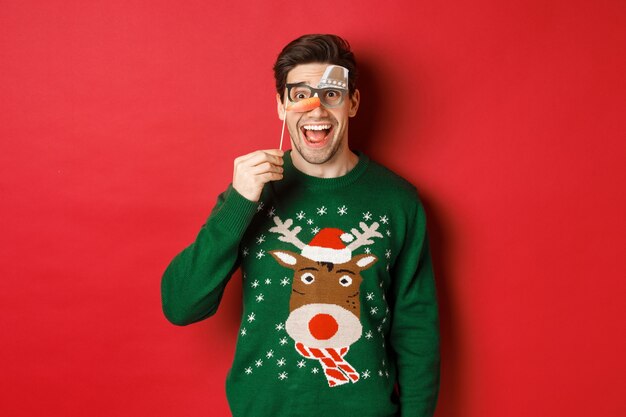 Портрет удивленного красавца в рождественском свитере, держащего забавную маску для вечеринки и улыбающегося, празднующего зимние праздники, стоящего на красном фоне