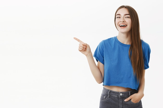 Портрет удивленной счастливой красивой женщины-брюнетки в модной синей укороченной футболке, указывающей влево и радостно смеющейся, весело обсуждающей забавное место для копирования или место, где можно повеситься над серой стеной