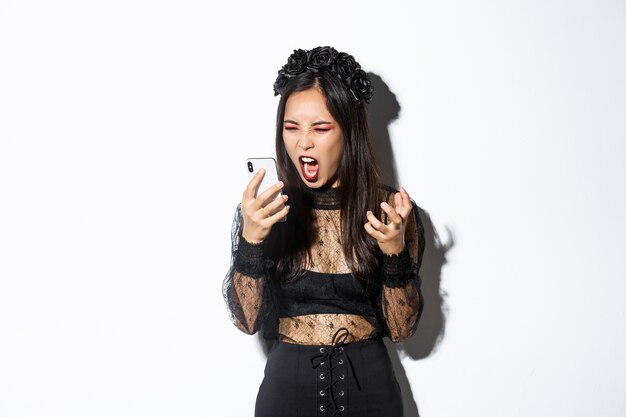 Portrait of angry asian woman in halloween costume looking mad, shouting at mobile phone and grimacing furious, standing over white background.