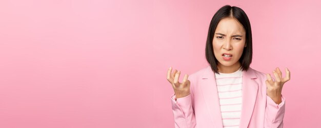 スーツの握りこぶしで怒っているアジアの女性の肖像画とピンクの背景の上に立っているsmthの悪い怒りに憤慨している