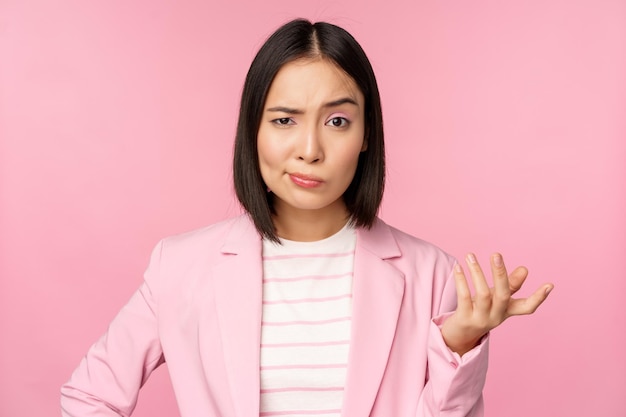 Портрет разгневанной азиатки в костюме, сжимающей кулаки и выглядящей в ярости, возмущенной чем-то плохим, стоящим на розовом фоне