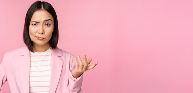 Portrait of angry asian woman in suit clench fists and looking furious outraged of smth bad standing over pink background