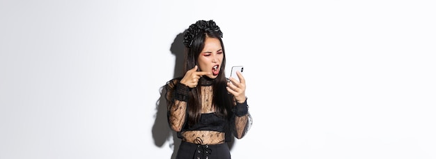 Portrait of angry asian woman in witch costume arguing on video call shouting at mobile phone and po