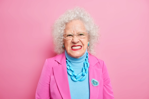 Portrait of angry curly haired senior woman squints face and looks unhappily, expresses negative emotions wears elegant costume with necklace and brooch red lipstick transparent glasses