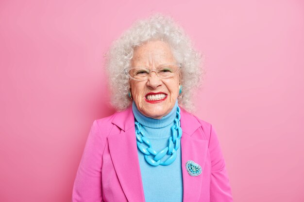 Portrait of angry curly haired senior woman squints face and looks unhappily, expresses negative emotions wears elegant costume with necklace and brooch red lipstick transparent glasses