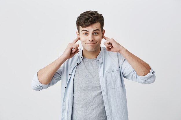 Portrait of angry and frustrated handsome man in blue shirt stopping his ears, plugging them with fingers, pursing lips while suffering from loud noise. Negative emotions. Stop this sound!