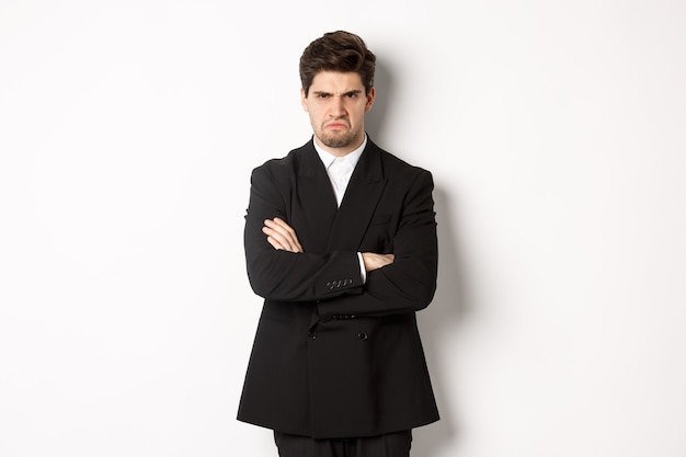 Portrait of angry handsome man in black suit, cross arms on chest and looking offended, frowning and pouting, being mad at someone, standing over white background