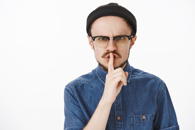 Portrait of angry irritated serious and strict male with beard in glasses and black beanie frowning shushing saying shh with index finger over mouth