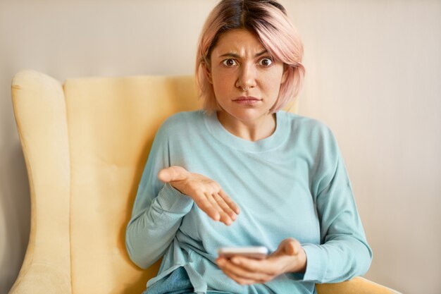 Portrait of angry irritated young Caucasian woman expressing negative emotions, holding smartphone