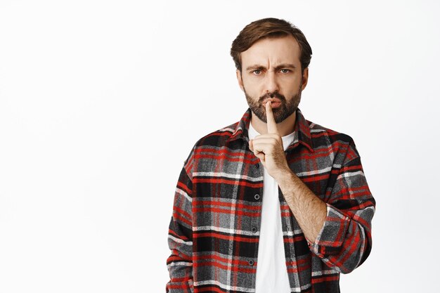 Portrait of angry man shows taboo gesture shushing making hush finger sign press forefinger to lips standing over white background