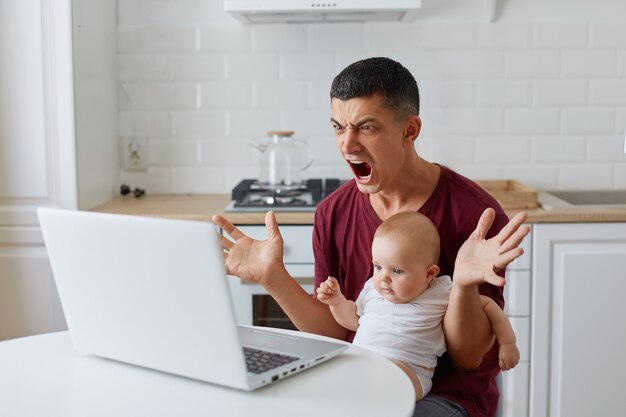 Portrait of angry man wearing maroon casual t shirt sitting at table with newborn baby boy or girl in kitchen in front of laptop, working online, having problems with project, screaming angrily.