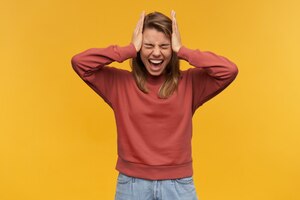 Portrait of angry screaming young woman in terra cotta sweatshirt with eyes closed and hands on head screaming and having a headache isolated over yellow wall