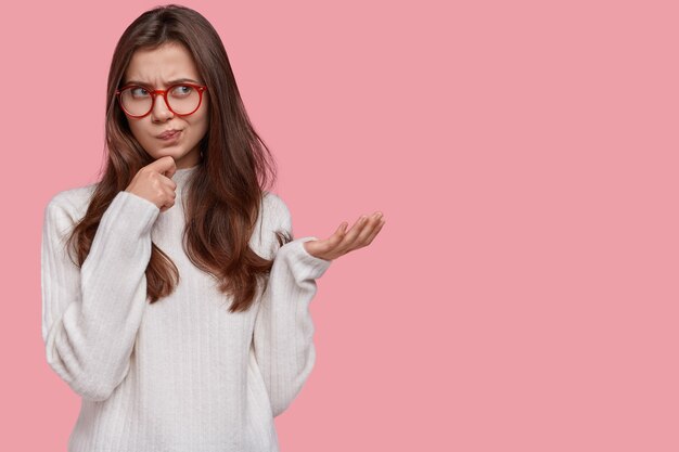 Portrait of angry woman holds hand under chin, purses lips and gestures angrily, wears white sweater, dissatisfied with price in shop, looks aside