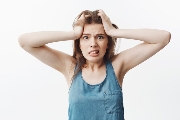 Portrait of angry young caucasian woman with dark hair in blue top squeezing head with hands,  with nervous face expression, trying to remember if she turned off iron.