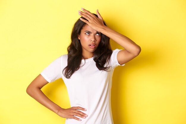 Portrait of annoyed african-american girl, slap forehead and roll her eyes bothered, forgot something important, standing over yellow background.