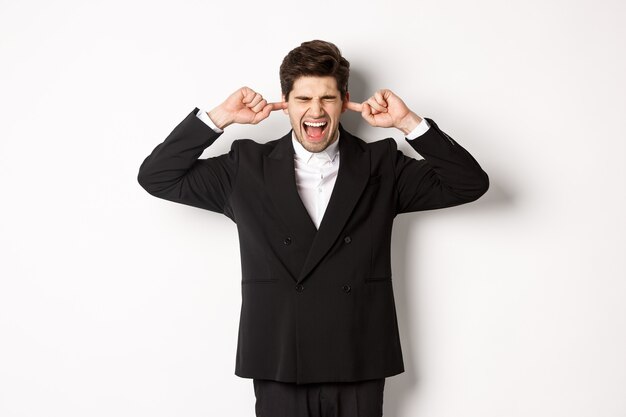 Portrait of annoyed and bothered businessman in black suit, shut ears and yelling, complaining loud noise, standing against white background.