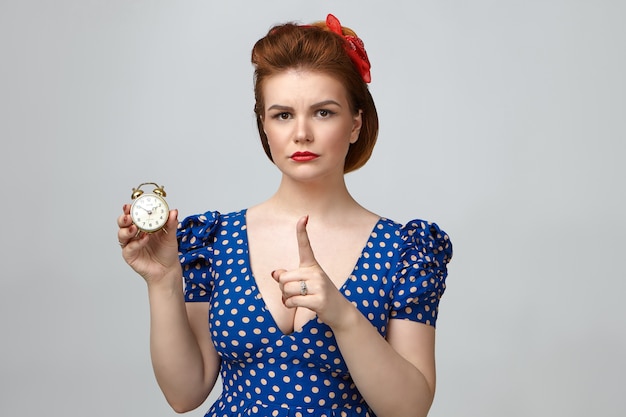 Portrait of annoyed frowning young mother wearing red lipstick, retro hairstyle and elegant dress with low cut neck holding vintage alarm clock, warning her teenage daughter to come home in time
