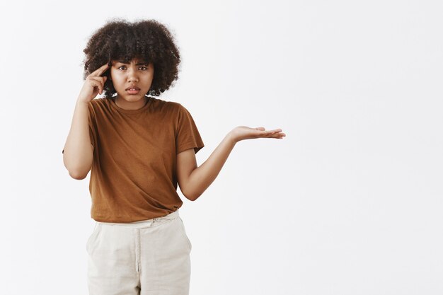 Portrait of annoyed and pissed questioned african american woman with afro hairstyle shrugging with spread palm and rolling index finger near temple scolding someone