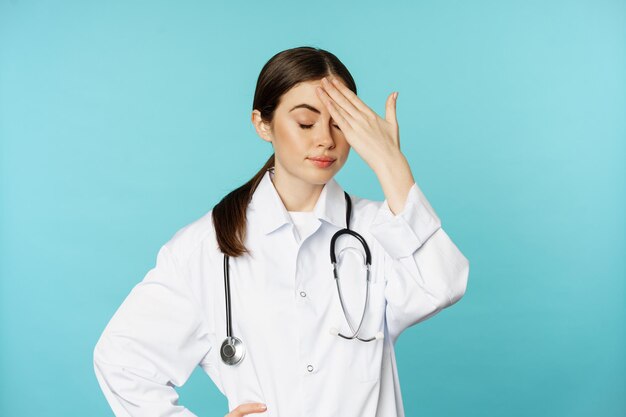 짜증나고 피곤한 여성 의사의 초상화, 얼굴 손바닥, 좌절된 눈, 멍청한 놈이 귀찮게 하고, 청록색 배경 위에 흰색 코트를 입고 서 있습니다.