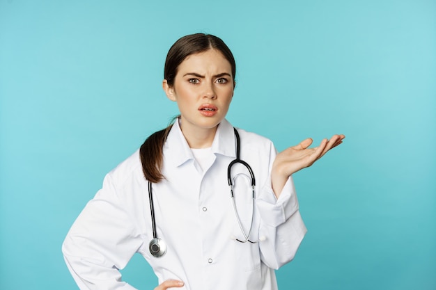 Portrait of annoyed, tired woman doctor, facepalm, roll eyes frustrated, bothered by smth stupid, standing in white coat over torquoise background.