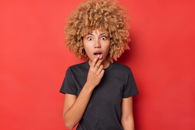 Portrait of anxious shocked woman with curly hair keeps jaw dropped cannot believe her eyes wears casual black t shirt feels frightened isolated over vivid red background hears something exciting