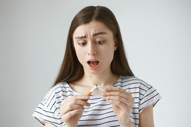 Portrait of anxious shocked young brunette female opening mouth in astonishment, having startled expression, looking at last cigarette that just broke in her hands, feeling panic and desperate