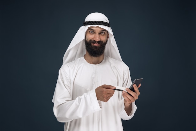 Foto gratuita ritratto di uomo d'affari arabo saudita su sfondo blu scuro per studio. uomo che utilizza smartphone per pagare la bolletta, acquisti online o scommesse. concetto di affari, finanza, espressione facciale, emozioni umane.