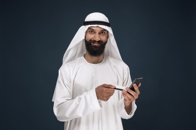 ダークブルーのスタジオの背景にアラビアのサウジアラビアの実業家の肖像画。請求書の支払い、オンラインショッピング、または賭けにスマートフォンを使用している男性。ビジネス、金融、顔の表情、人間の感情の概念。